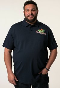 Námořnicky modré polo tričko vyrobené z bavlny, s barevným vyšíváním "Exotický ostrov" na levém prsu. Krátké rukávy a límec.