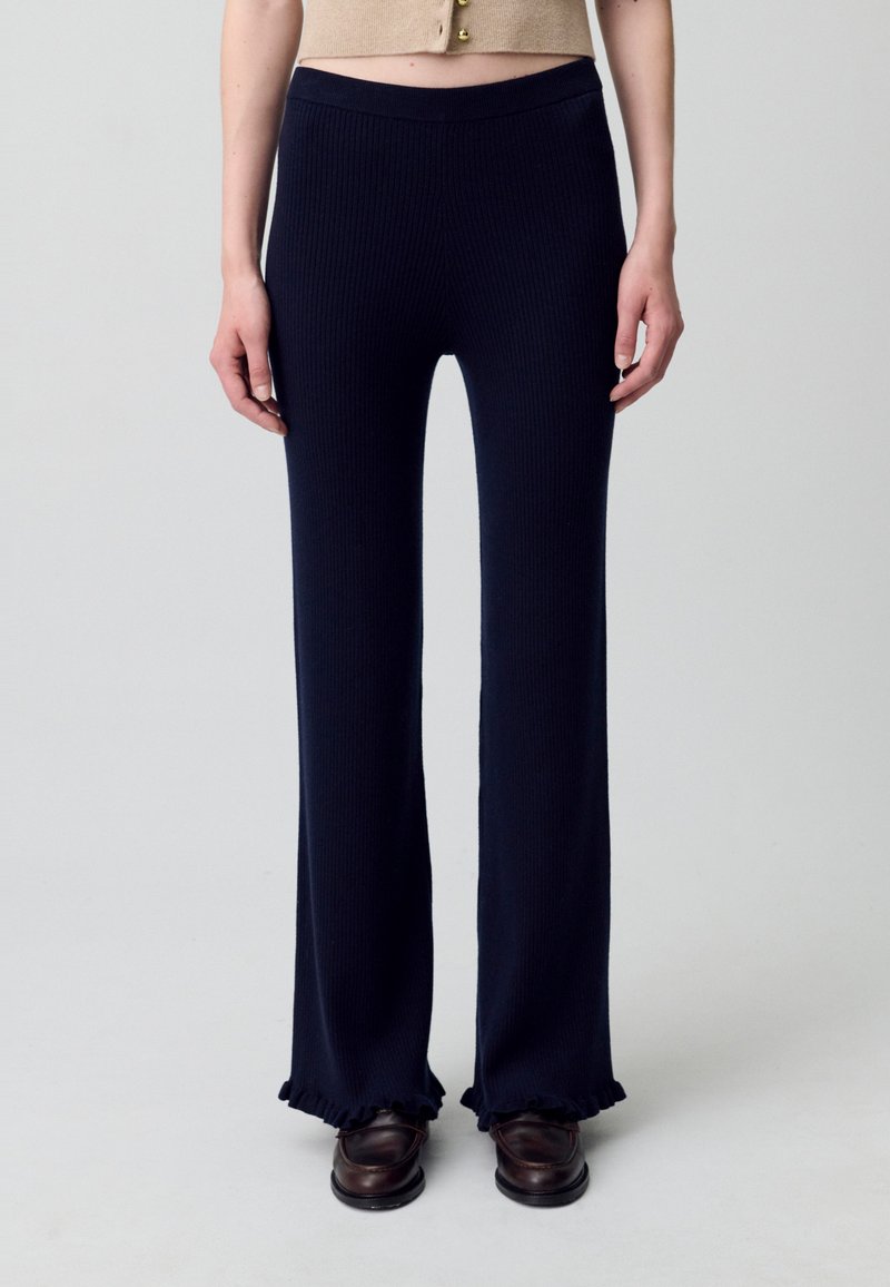 Pantalones de punto acanalado en un azul marino oscuro con perneras acampanadas y dobladillos con volantes, que presentan una cinturilla lisa. Se llevan con zapatos de cuero marrón.