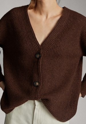 Vrouw draagt een bruine geribbelde gebreide cardigan met twee grote knopen, gecombineerd met gebroken witte broek, handen in de zij, zichtbare nek en sleutelbeen.