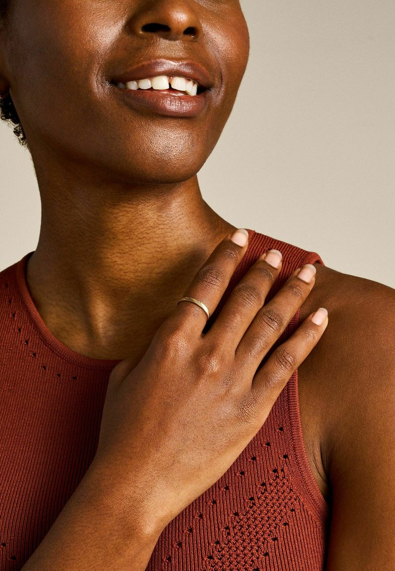 Gold ring with a twisted design, worn on the index finger. Hand resting on a rust-colored, ribbed tank top with small holes.