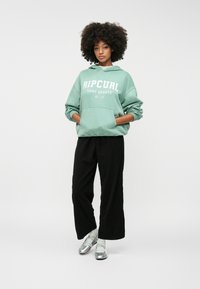 Mintgroene oversized hoodie met wit "Ripcurl Surf Sports" logo, gecombineerd met zwarte wijdpijpjurken en zilveren schoenen.