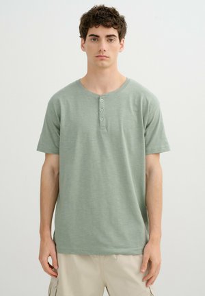 Joven con cabello rizado que lleva una camiseta Henley de manga corta en verde salvia y pantalones cargo beige, de pie sobre un fondo blanco liso.