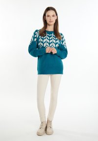 Pulover teal cu model alb de frunze pe mâneci, guler rotund și croială lejeră. Asortat cu pantaloni ajustați de culoare crem și cizme bej cu șireturi.