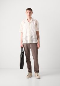 Cămașă cu mânecă scurtă, de culoare albă-crem, cu un model subtil, asortată cu pantaloni maro și adidași bej, ținând o geantă de umăr neagră.