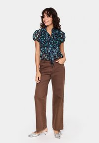 Prozoren črn top z modrim in zelenim tiskom perja, naborki na rokavih in zaveza na izrezu, v kombinaciji z visokim pasom rjavih hlač.