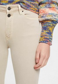 Jeans ajustados beige de canalé con dos botones negros y un bolsillo frontal, combinados con un suéter de punto multicolor con mangas estampadas.