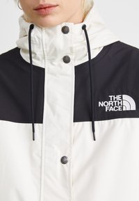 Veste résistante aux intempéries blanche et noire avec un col haut, un cordon de serrage ajustable et un logo de marque sur la poitrine. Comprend des fermetures à boutons.