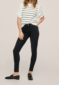 Jeans negros ajustados con una cintura alta, combinados con un chaleco de punto blanco a rayas y mocasines negros. Textura suave, diseño ceñido.