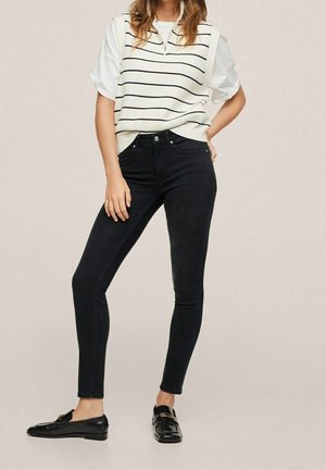 Jeans negros ajustados con una cintura alta, combinados con un chaleco de punto blanco a rayas y mocasines negros. Textura suave, diseño ceñido.