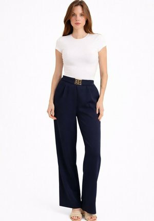Femme debout, portant un haut blanc ajusté à manches courtes, un pantalon large bleu marine taille haute avec une ceinture décorative et des sandales beige.