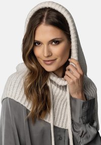 Indumento a cappuccio in maglia color crema con texture a coste, abbinato a una camicia grigia con bottoni, caratterizzata da maniche lunghe e polsini arricciati.
