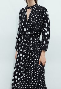 Schwarzes Wickelkleid mit weißen geometrischen Mustern, langen Ärmeln, einem Bindegürtel in der Taille und einem Schlüssel-Ausschnitt. Glatte Stofftextur.