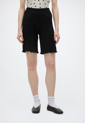 Personne portant un short noir côtelé jusqu'aux genoux, des chaussettes blanches, des ballerines noires et un haut blanc sans manches à pois noirs, debout sur un fond blanc.