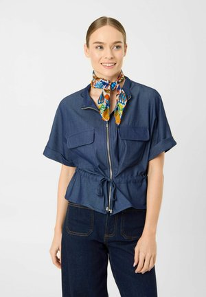 Vrouw met een blauw kortemouws shirt met ritssluiting en trekkoord in de taille, donkere spijkerbroek en een kleurrijke bloemenhalsdoek, glimlacht en kijkt naar rechts.