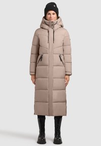 Hosszú, bézs pufi kabát kapucnival és elöl cipzárral. Cipzáras zsebekkel és steppelt mintával rendelkezik. Fekete sapkával és katonai bakancsokkal viselik.