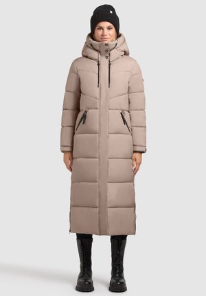 Lang beige dynejakke med hætte og lynlås foran. Har lynlåslommer og et quiltet design. Båret med en sort hue og kampstøvler.