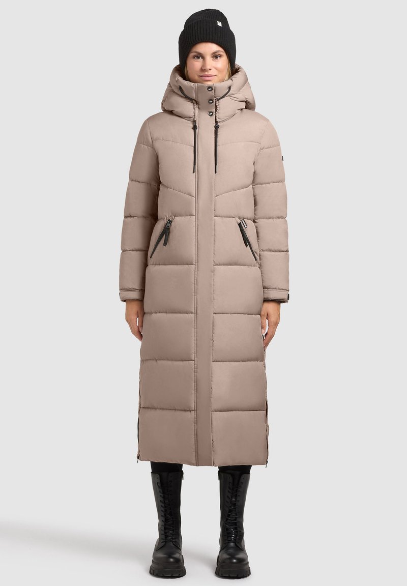 Hosszú, bézs pufi kabát kapucnival és elöl cipzárral. Cipzáras zsebekkel és steppelt mintával rendelkezik. Fekete sapkával és katonai bakancsokkal viselik.