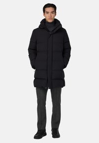 Pikk must puffjakker koos kapuutsi ja esise paljandiga, mis sisaldab lukku ja nööpe, mis on horisontaalse õmbluskujundusega. Kantud hallide pükste ja mustade kingade kohal.