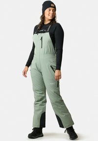 Gröna isolerade overaller med justerbara remmar, framficka med dragkedja, sidodragkedjor och svarta detaljer vid anklarna. Texturerat tyg för värme.