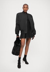 Giacca bomber nera con polsini e colletto a costine, abbinata a un vestito nero, collant neri trasparenti e scarpe con tacco. Accessoriata con una borsa di pelliccia nera.