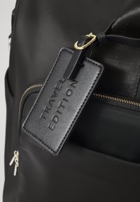 Fekete utazótáska texturált felülettel, cipzáras zsebbel, arany színű kiegészítőkkel és egy téglalap alakú bőrcímkével, amelyen "UTAZÁS KIADÁS" felirat olvasható.