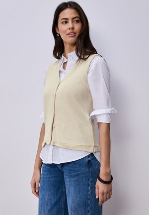 Vrouw draagt een beige gebreid vest met knopen over een wit shirt met opgerolde mouwen en blauwe spijkerbroek, staand tegen een effen lichte achtergrond.