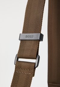 Brun stofrem med et struktureret væv; har en sort plastikspænde mærket "BOSS." Bemærkelsesværdig for sin solide konstruktion og justerbare længde.