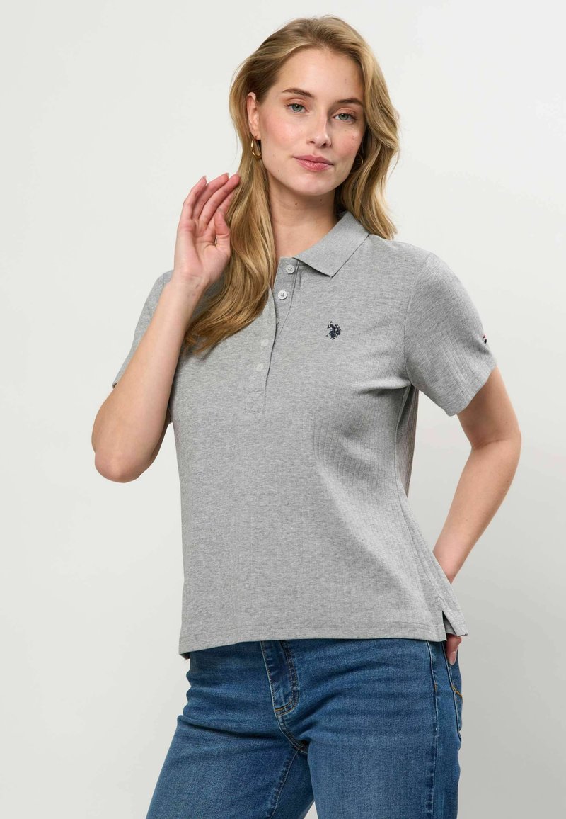 Femme aux longs cheveux blonds portant un polo gris clair à manches courtes et un jean bleu, posant avec une main près de son visage.