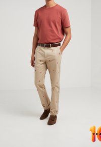 Uomo che indossa una maglietta color ruggine, pantaloni beige con piccoli disegni ricamati rossi, cintura marrone e mocassini in suede marrone, in piedi con le mani nelle tasche.