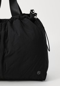 Schwarze Stofftasche mit gerafft oben, glatter Textur, verstellbarem schwarzen Tragegurt und einem silbernen Logodetail an der Seite.