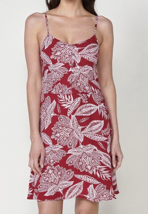 Femme portant une robe rouge sans manches avec des motifs floraux blancs et des bretelles fines, debout devant un fond uni.