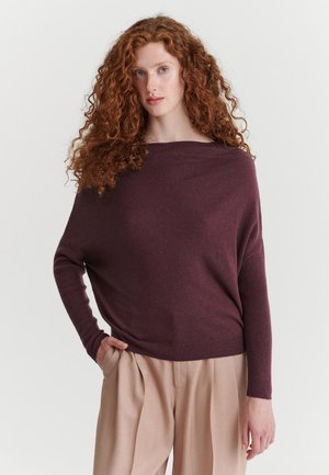 Mujer con largo cabello rizado y rojo, vestida con un suéter holgado de color púrpura oscuro y pantalones beige, de pie con una mano en el bolsillo frente a un fondo sencillo.