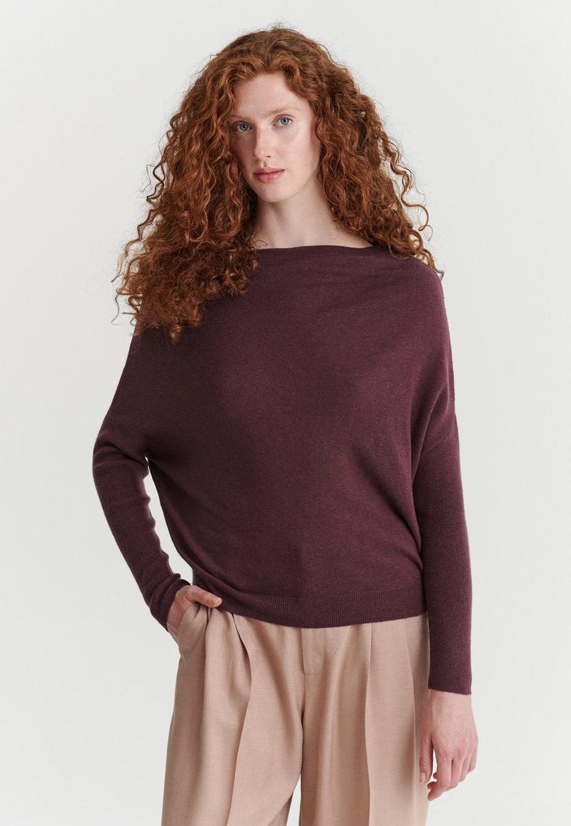 Femme aux longs cheveux roux bouclés portant un pull ample violet foncé et un pantalon beige, debout avec une main dans la poche devant un fond uni.