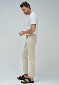 Pantalones de algodón beige con un corte slim, que incorporan bolsillos laterales, cierre de botón y trabillas para cinturón. Combinados con una camiseta blanca de cuello redondo y sandalias negras.