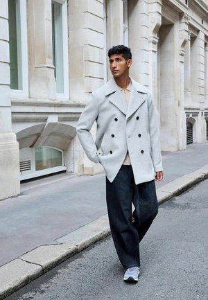 Light grey double-breasted coat with large black buttons, paired with loose black pants and grey sneakers, standing on a city street.