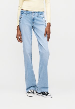 Ανοιχτό μπλε τζιν καμπάνα με ψηλή μέση και πλευρικές λεπτομέρειες με κουμπιά, συνδυασμένα με μαύρα και λευκά αθλητικά παπούτσια και ανοιχτό κίτρινο μακρυμάνικο τοπ.