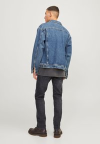 Casaco de ganga azul com um corte descontraído, apresentando um colarinho, bolsos laterais e punhos com botões. Usado sobre uma camisa cinza e jeans pretos.