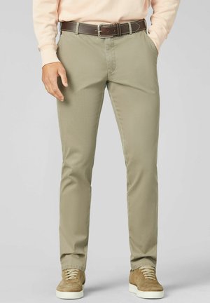 Mann trägt beige Chinohosen, einen braunen Ledergürtel, ein hellbeiges langärmeliges Hemd und beige Wildleder-Sneaker, steht mit einer Hand in der Tasche.