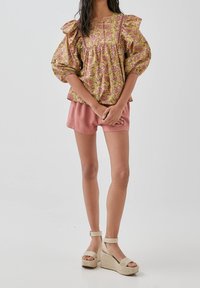 Femme portant un chemisier à manches bouffantes à fleurs, un short rose poudré et des sandales à plateforme à talons hauts beige, se tenant les mains jointes.
