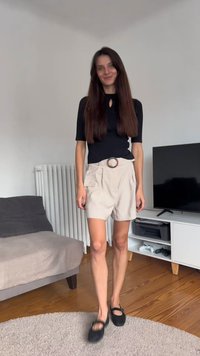 Čierny krátky rukáv top s výrezom v tvare kľúčovej dierky, béžové šortky s opaskom a bočnými vreckami, čierne klumpačky, stojaci v obývacej izbe.