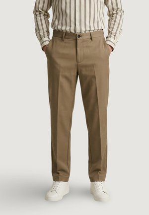 Mężczyzna ubrany w beżowe proste spodnie z przednią zaszewką, w paskowaną koszulę z długim rękawem wsuniętą w spodnie oraz białe trampki, ręce w kieszeniach.