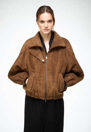 Kobieta nosząca brązową, oversize'ową kurtkę zamszową z elastycznymi mankietami i ściągaczem u dołu, stojąca z rękami w kieszeniach na tle jasnego, jednolitego tła.