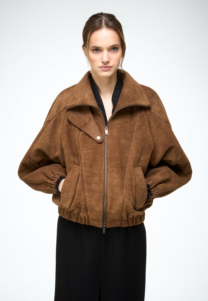 Žena v hnedej nadrozmernej semišovej bunde s elastickými manžetami a lemom, stojaca s rukami v vreckách pred jednoduchým svetlým pozadím.