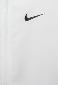 Hvid sportsjakke lavet af glat stof, med et sort Nike swoosh-logo. Lynlåslukning der løber langs forsiden. Minimalistisk design.