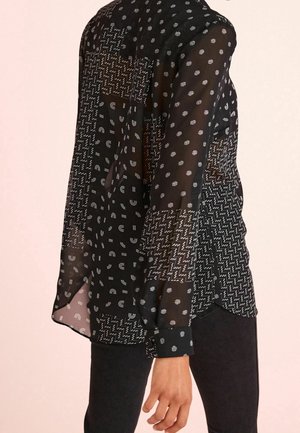 Blouse noire en voile transparent avec divers motifs géométriques, y compris des points et des zigzags. Manches longues avec détails aux poignets, ourlet courbé.