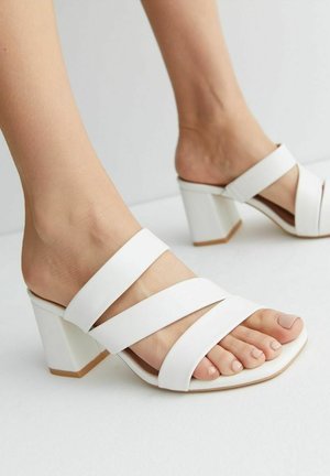 Białe klapki na obcasie mają szerokie skrzyżowane paski, kwadratowy czubek i masywny obcas. Materiał wydaje się gładki z matowym wykończeniem.