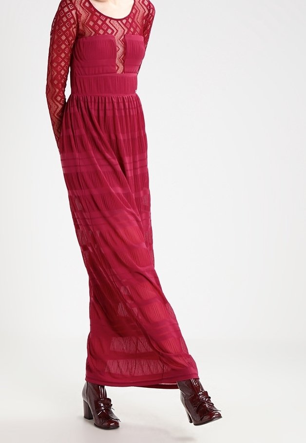Femme portant une longue robe rouge transparente avec des manches en dentelle et des bottes à talons rouge foncé, debout devant un fond clair uni.