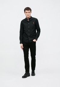 Camisa preta com botões e gola, com um bolso e etiqueta castanha, combinada com calças justas pretas e botas pretas. Fundo liso.