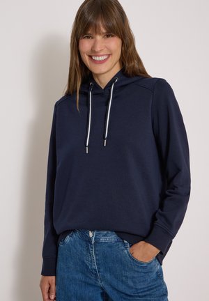 Femme souriante aux cheveux bruns porte un sweat à capuche bleu marine avec des cordons blancs et un jean bleu, debout avec une main dans la poche.