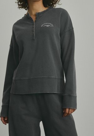 Persoon die een donkergrijs henley-shirt met lange mouwen draagt met de tekst "New York City, NY" en bijpassende broek tegen een effen achtergrond.